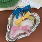 A heart-shaped silver dish holding three resin bow-shaped hair clips in blue, yellow, and pink.