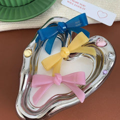 A heart-shaped silver dish holding three resin bow-shaped hair clips in blue, yellow, and pink.