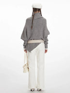 Woman wearing a grey asymmetrical knit poncho with a turtleneck, white pants, and a white belt, back view