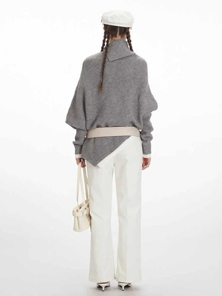 Woman wearing a grey asymmetrical knit poncho with a turtleneck, white pants, and a white belt, back view
