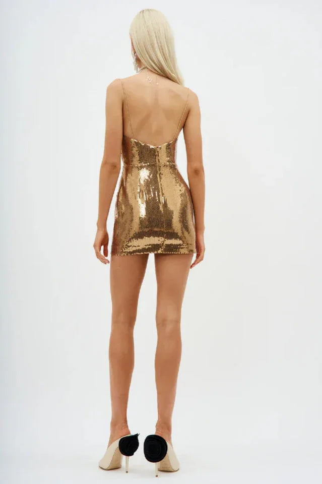 Back view of a woman wearing a gold sequined mini dress with a cowl neckline and spaghetti straps, standing against a white background.