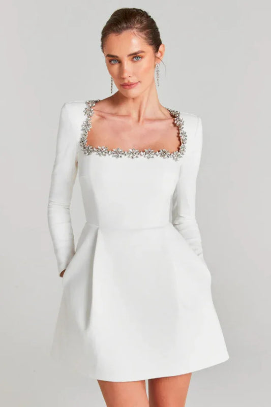 Woman wearing a white long-sleeve mini dress with a square neckline adorned with floral embellishments and a pleated skirt.