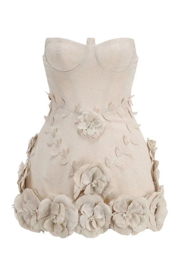 Full front view of a strapless beige mini dress with a fitted corset-style bodice. The skirt is decorated with appliqué flowers and leaves, with larger floral embellishments at the hem.