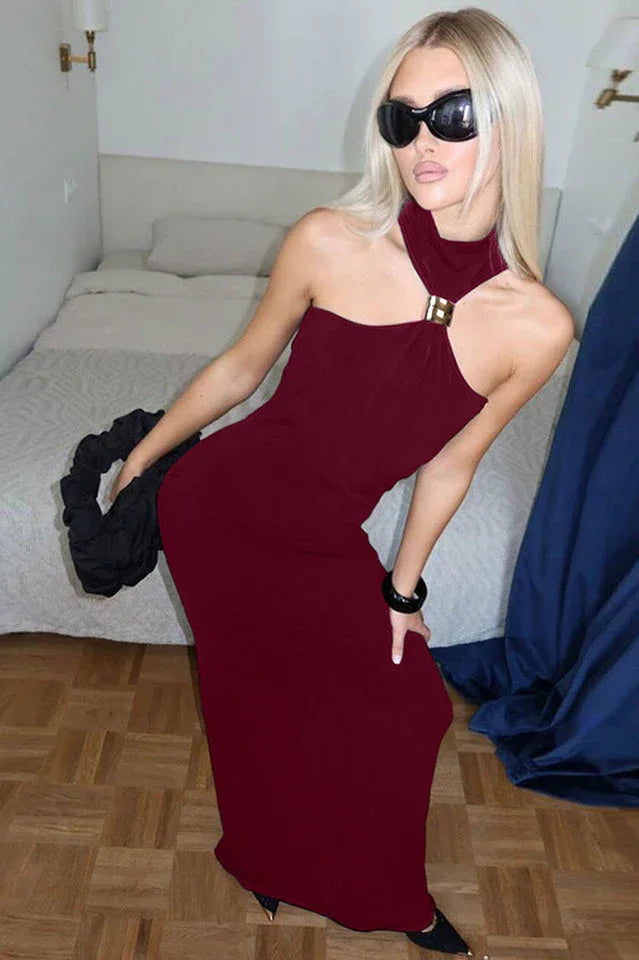 Woman with blonde hair wearing a burgundy sleeveless halter maxi dress with a gold-tone hardware detail at the neckline and sunglasses, posing in a bedroom.