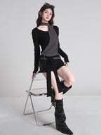 Full-length shot of a woman wearing a black long-sleeved top with a grey asymmetrical overlay and scarf detail, black shorts, and black boots, sitting on a chair.