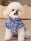 A fluffy white dog wearing a blue plaid dog vest with white buttons and ruffled sleeves, sitting indoors.