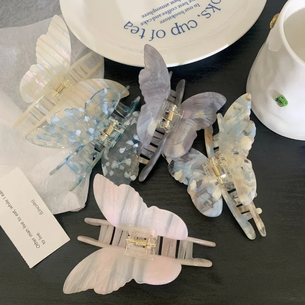 A collection of butterfly-shaped hair claw clips in various pastel colors, including blue, purple, pink, and white, arranged on a dark surface.