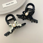 Two black hair claw clips with pearl accents, one with a black bow and one with a light green bow, on a plate
