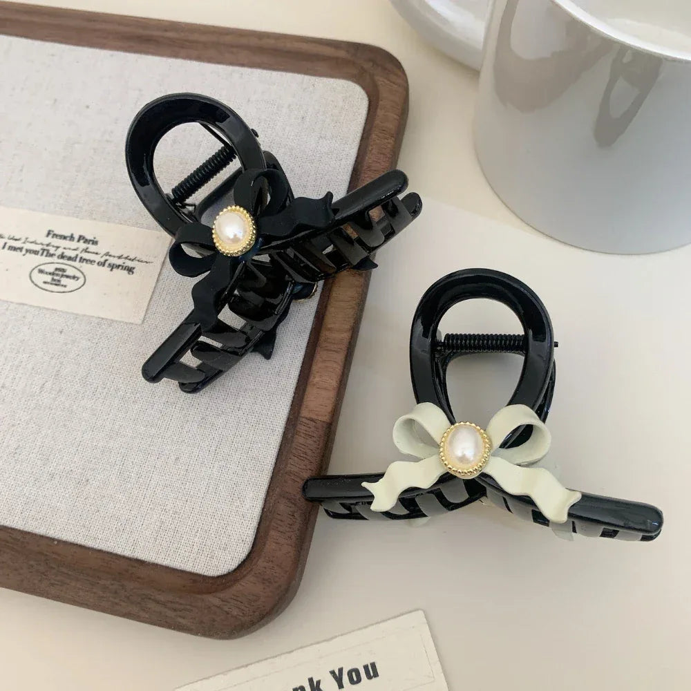 Two black hair claw clips with pearl accents and bows, placed on a tray with a mug in the background