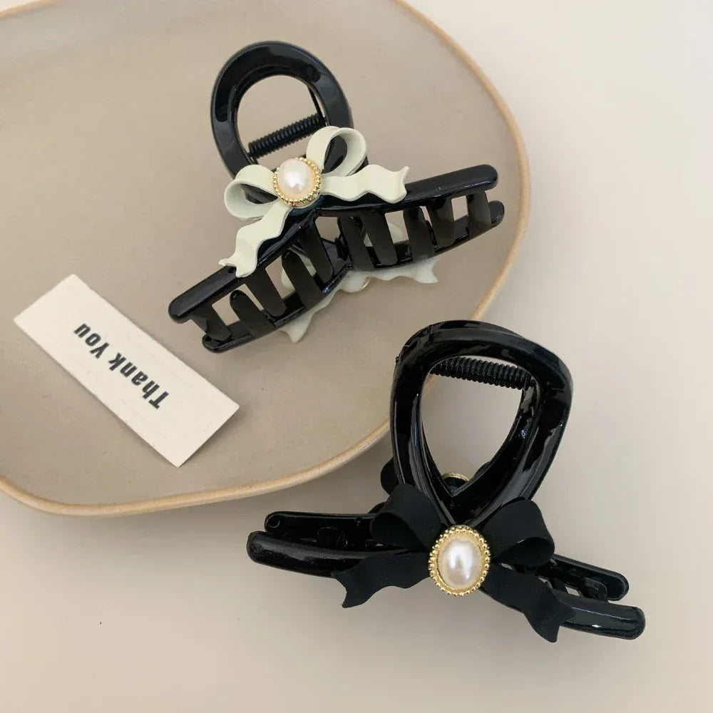 Two black hair claw clips with pearl accents, one with a black bow and one with a light green bow, on a plate