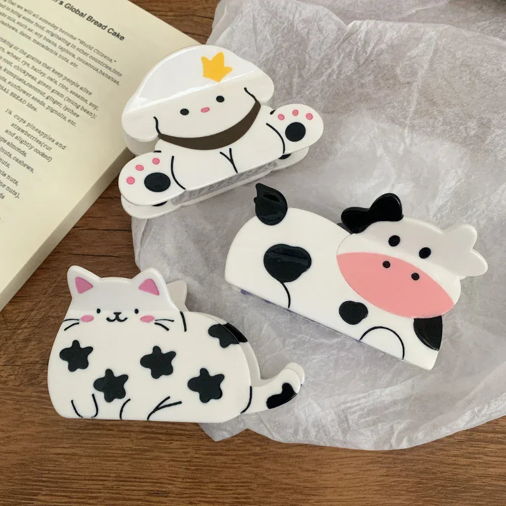 A variety of cute animal hair clips, including a cow, a dog, and a cat, are arranged on a white surface next to an open book.