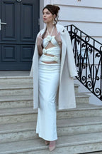 Woman wearing a white satin halter maxi dress with cut-out detailing and floral embellishments, with a cream coat draped over her shoulders, standing on stairs
