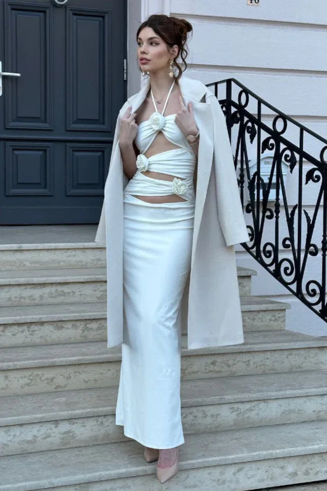 Woman wearing a white satin halter maxi dress with cut-out detailing and floral embellishments, with a cream coat draped over her shoulders, standing on stairs