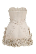 Back view of a strapless beige mini dress with a fitted bodice and a full skirt. The back of the bodice has a zipper closure and the skirt is heavily embellished with fabric flowers.