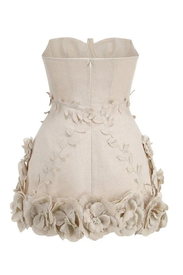 Back view of a strapless beige mini dress with a fitted bodice and a full skirt. The back of the bodice has a zipper closure and the skirt is heavily embellished with fabric flowers.