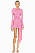 Model wearing a pink long-sleeve mini dress with asymmetrical draping and a twist detail at the neckline. The dress has a cutout detail at the chest and a ring hardware detail on the hip. The hem is asymmetrical with a draped panel. She is holding a silver clutch.