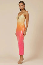 Side profile of a model wearing a yellow, orange, and pink ombre sequined midi dress with spaghetti straps and a star embellishment at the bust, against a neutral background.