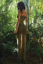 Back view of a woman wearing a strapless, shimmering olive green gown with ruched detailing and a knotted front accent, standing in a lush green environment.
