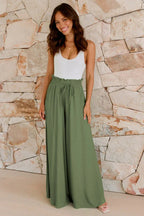 Woman wearing a white tank top and olive green wide-leg palazzo pants standing against a stone wall.