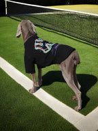 Weimaraner dog wearing a black t-shirt with a colorful graphic print and silver chain, standing on green turf near a tennis net, showing the back of the shirt.