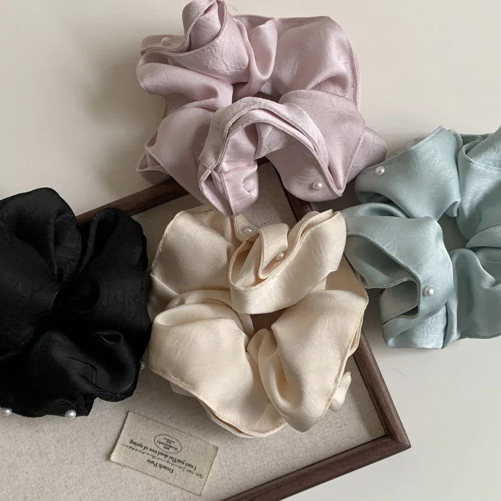 A collection of four scrunchies in pastel pink, light blue, cream, and black, each adorned with small pearls, arranged on a white surface with a wooden tray and a tag.
