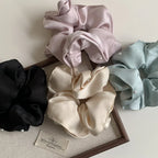 A collection of four scrunchies in pastel pink, light blue, cream, and black, each adorned with small pearls, arranged on a white surface with a wooden tray and a tag.