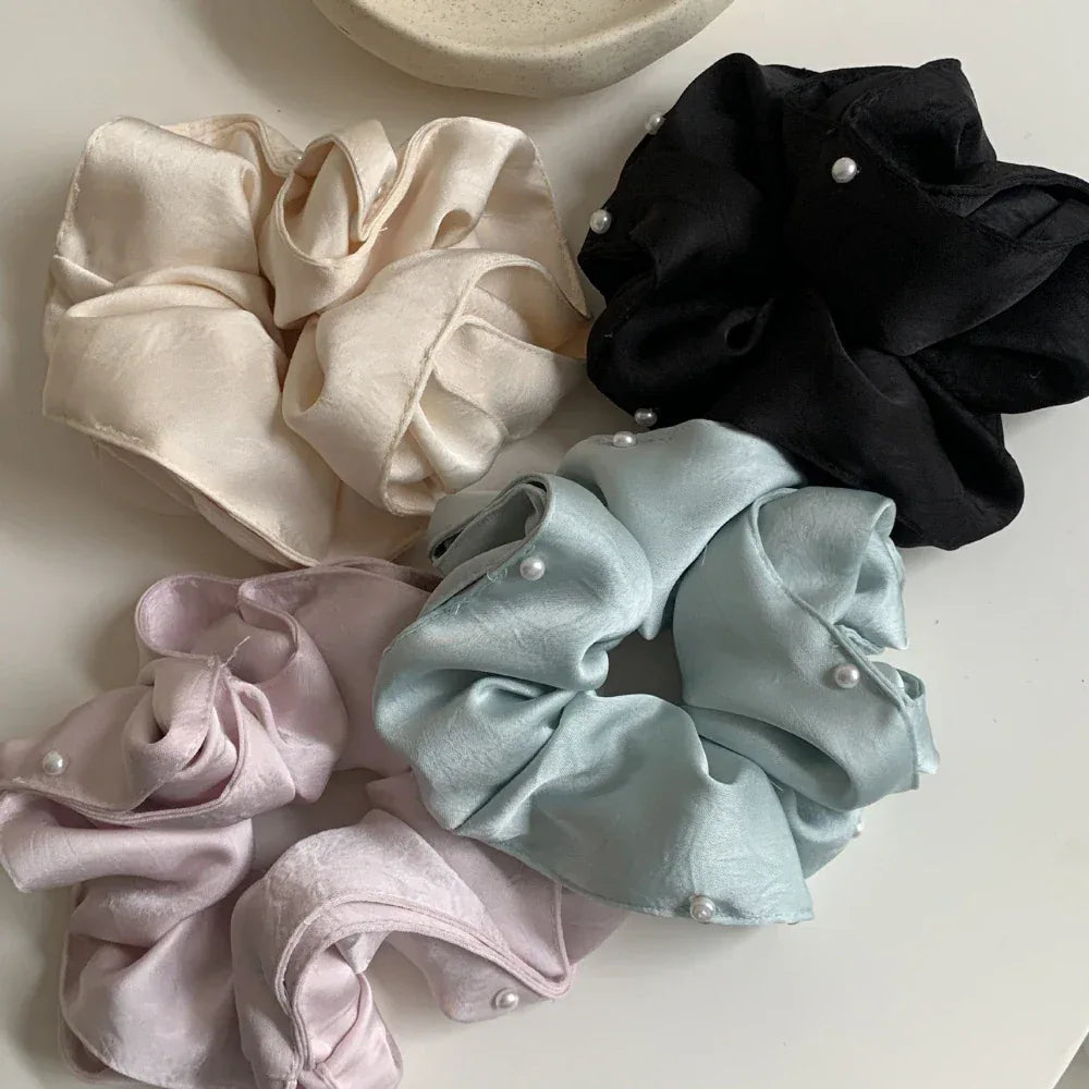 Top-down view of four scrunchies in cream, black, light blue, and pastel pink, all decorated with pearls, laid out on a white surface.
