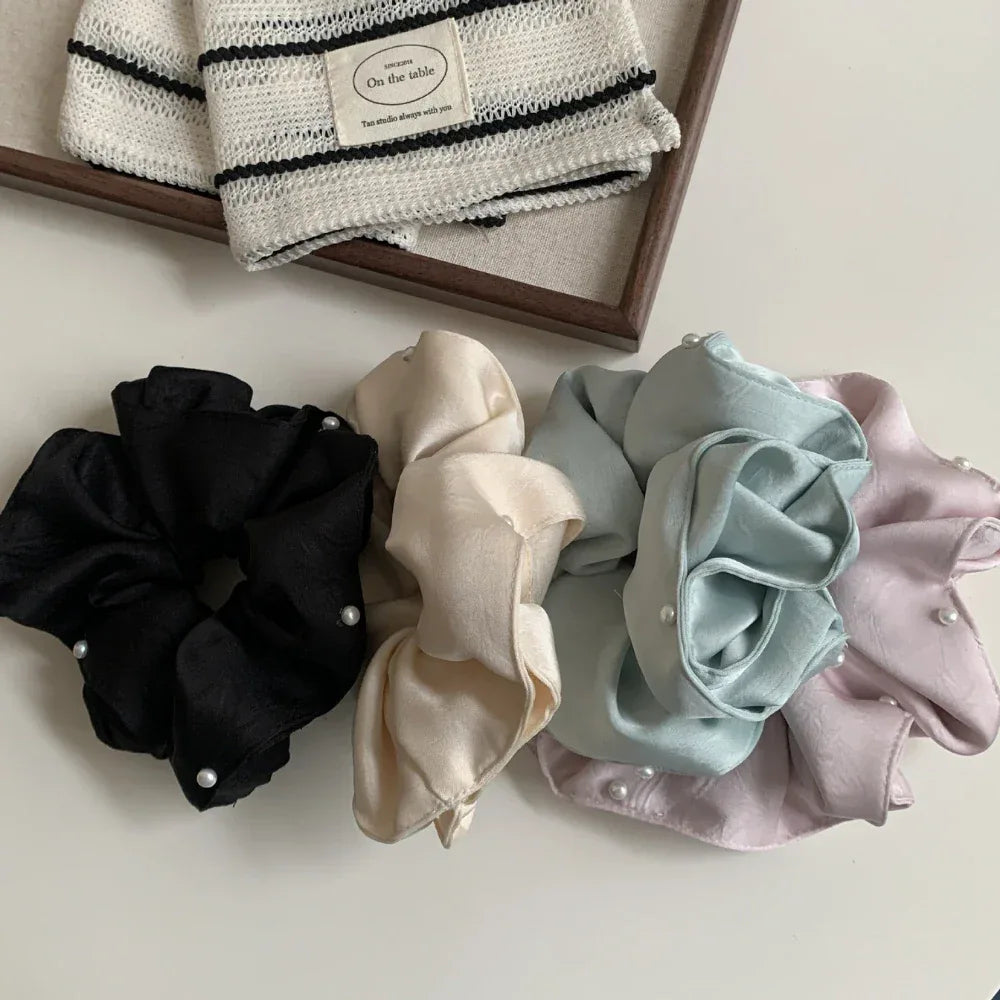 Four scrunchies in black, cream, light blue, and pastel pink, each with pearl accents, are displayed on a white surface next to a wooden tray containing a striped fabric item.