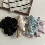 Four scrunchies in black, cream, light blue, and pastel pink, each with pearl accents, are displayed on a white surface next to a wooden tray containing a striped fabric item.