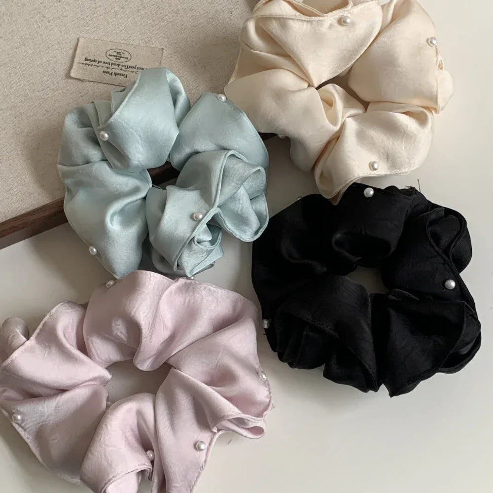 Overhead view of four scrunchies: cream, light blue, pastel pink, and black. Each scrunchie features scattered pearl embellishments and is arranged on a white surface.