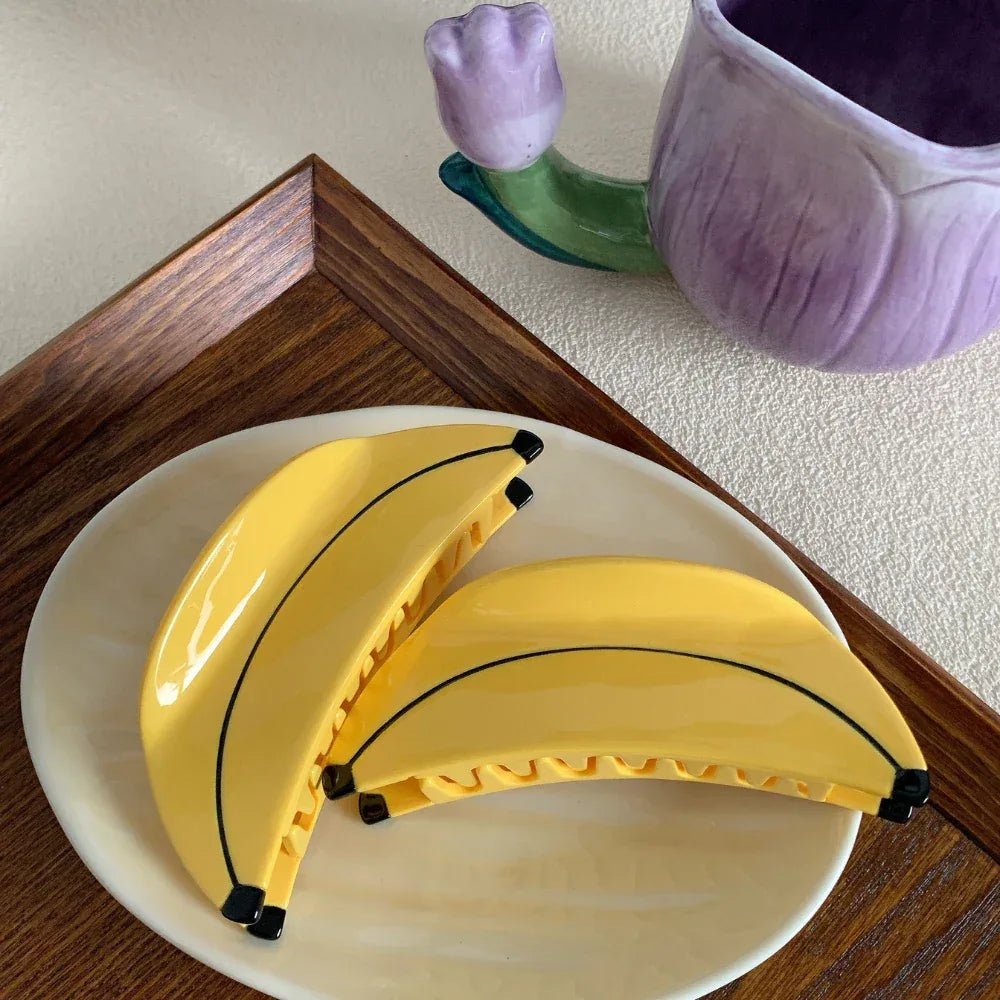 Two yellow banana-shaped hair clips resting on a cream-colored plate, with a wooden tray and a purple floral mug in the background.