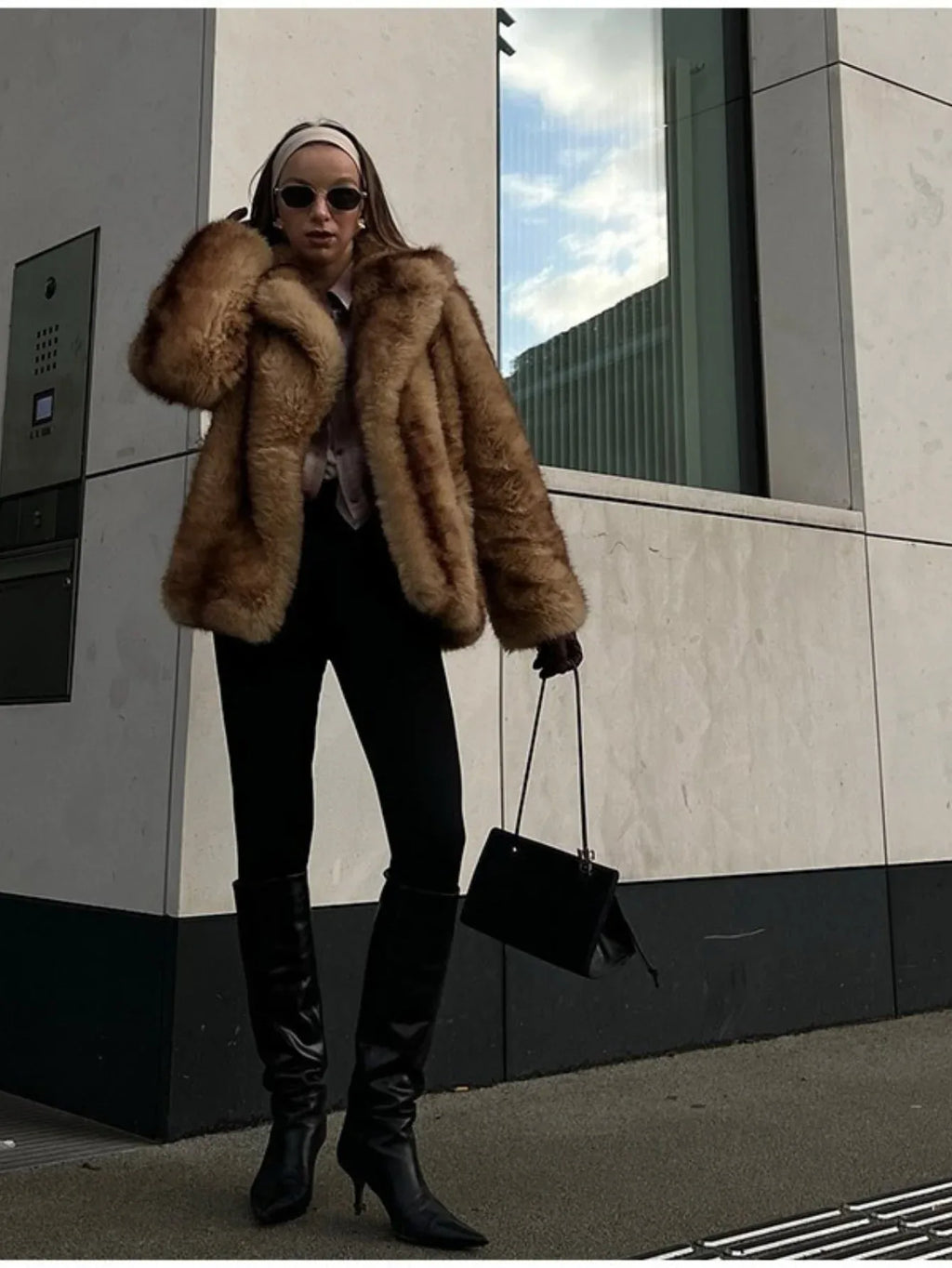 Woman wearing a tan and brown faux fur cropped jacket with black pants and black boots, holding a black handbag.