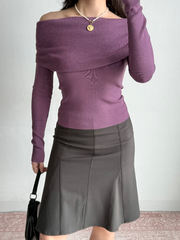 Woman wearing a purple off-the-shoulder knit top with a draped neckline, long sleeves, a pearl necklace, and a dark grey skirt, holding a black handbag.