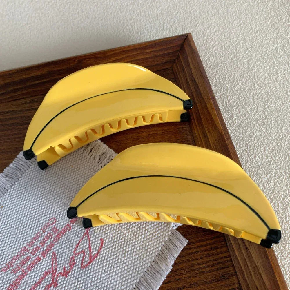 Two yellow banana-shaped hair clips positioned on a textured white fabric with a wooden tray in the background.