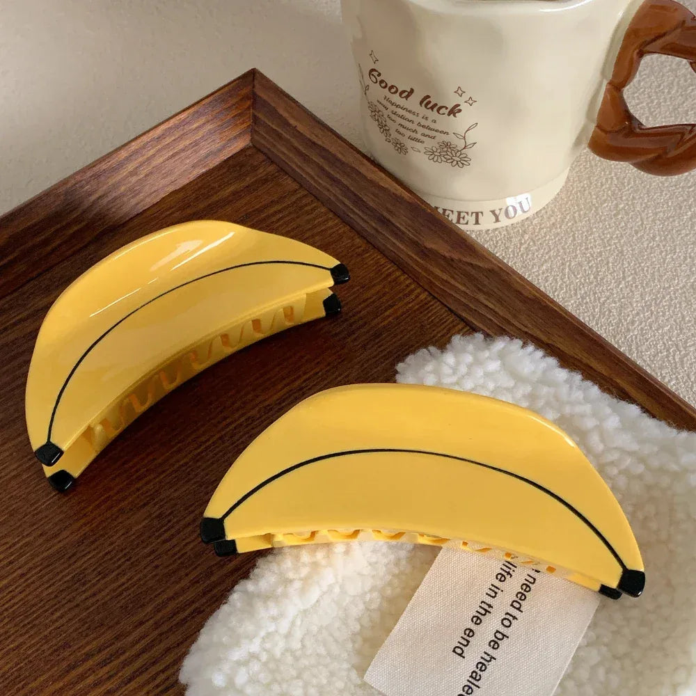 Two yellow banana-shaped hair clips on a fluffy white surface with a tag, next to a wooden tray and a mug.