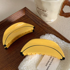 Two yellow banana-shaped hair clips on a fluffy white surface with a tag, next to a wooden tray and a mug.
