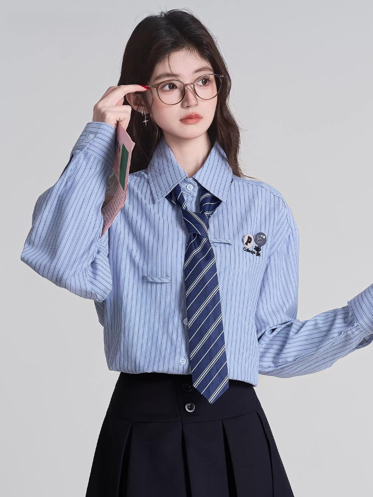 Close-up of a model wearing a light blue striped button-down shirt with a navy striped tie and a black pleated skirt, holding a pink wallet.