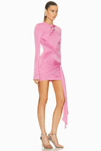 Side view of a model wearing a pink long-sleeve mini dress with asymmetrical draping and a twist detail at the neckline. The dress has a cutout detail at the chest and a ring hardware detail on the hip. The hem is asymmetrical with a draped panel.