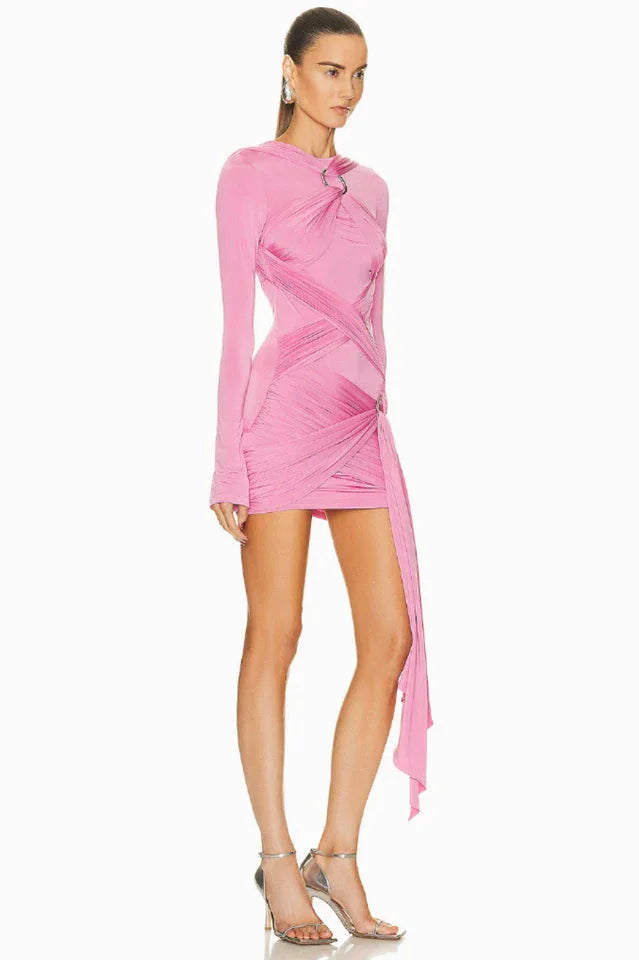 Side view of a model wearing a pink long-sleeve mini dress with asymmetrical draping and a twist detail at the neckline. The dress has a cutout detail at the chest and a ring hardware detail on the hip. The hem is asymmetrical with a draped panel.