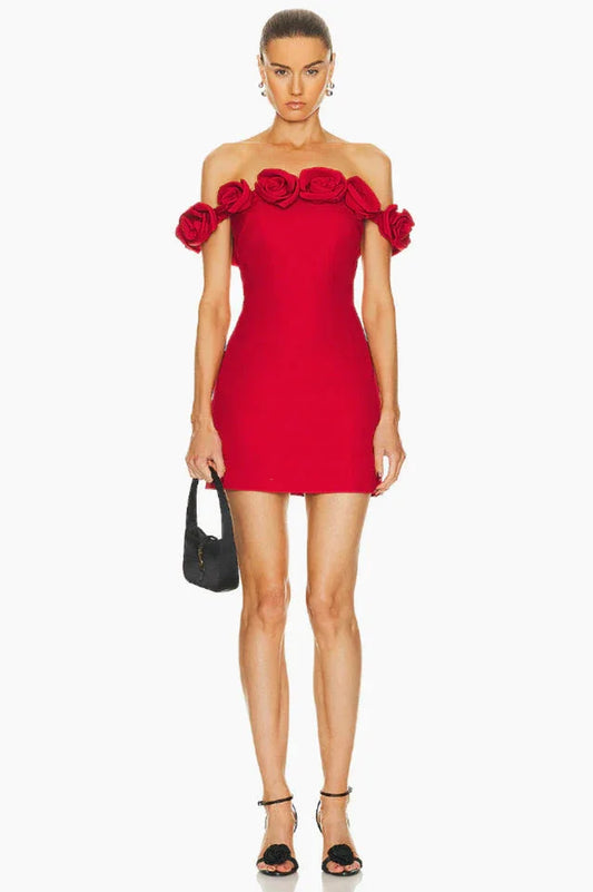 Woman wearing a red off-the-shoulder mini dress with rose embellishments on the neckline, holding a black clutch and wearing black strappy heels.