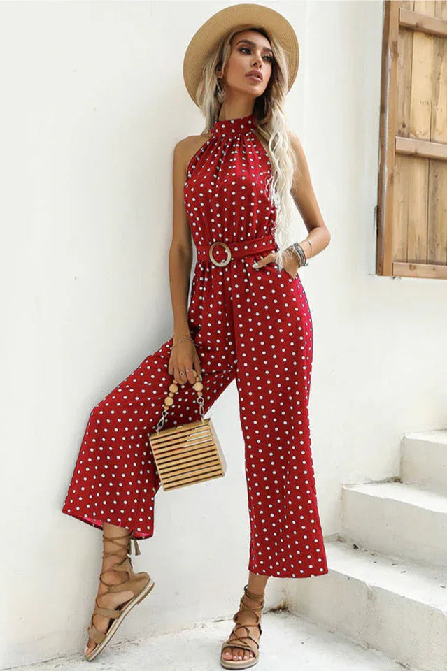 Woman wearing a red polka dot halter neck wide leg jumpsuit with a belt, straw hat, and wooden handbag.