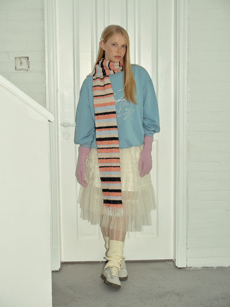 A woman stands wearing a light blue sweatshirt, a white tutu skirt, pink gloves, leg warmers, sneakers, and a striped scarf.