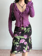 Full body shot of a woman wearing a purple floral lace long-sleeve top, floral skirt, and a black handbag.