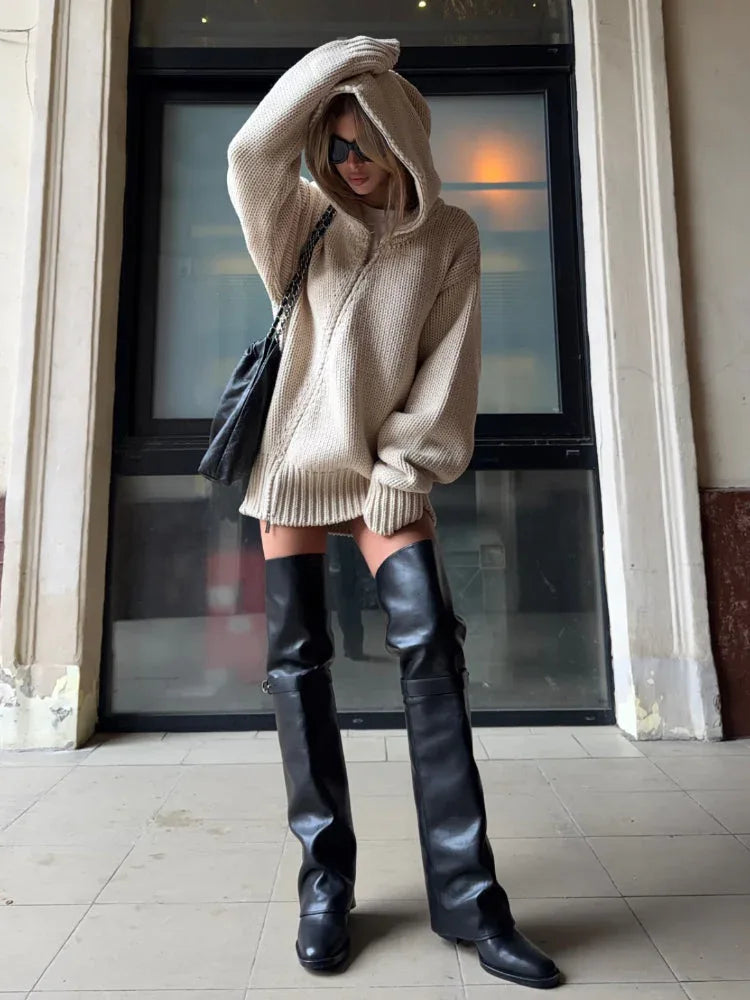 Woman wearing a beige oversized knit hooded sweater dress and black thigh-high boots, adjusting her hood.