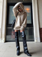 Woman wearing a beige oversized knit hooded sweater dress and black thigh-high boots, adjusting her hood.