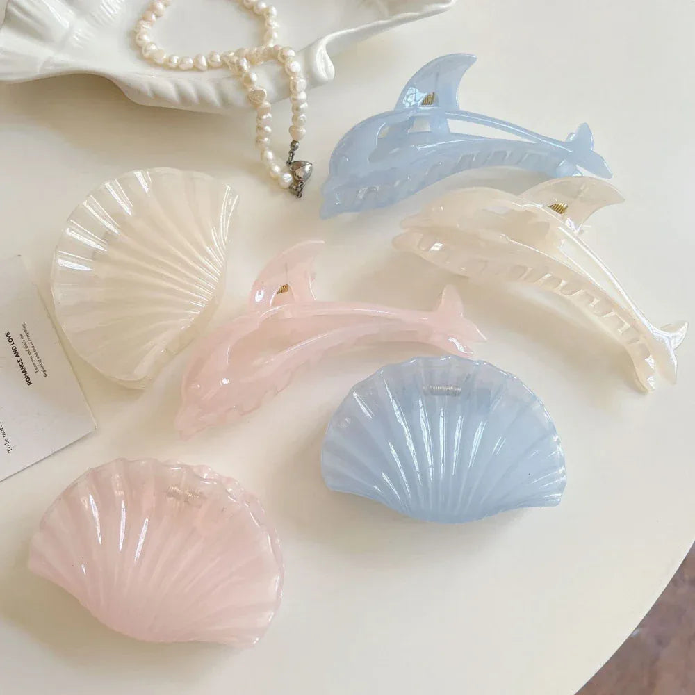 Top-down view of pastel dolphin and seashell hair claw clips with a pearl necklace and a card on a white surface