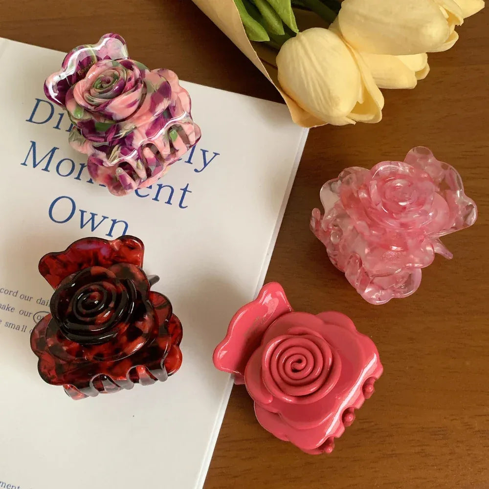 Four rose-shaped hair claw clips in various colors including pink, purple, and red tortoise shell pattern, arranged on a book and a wooden surface with yellow flowers