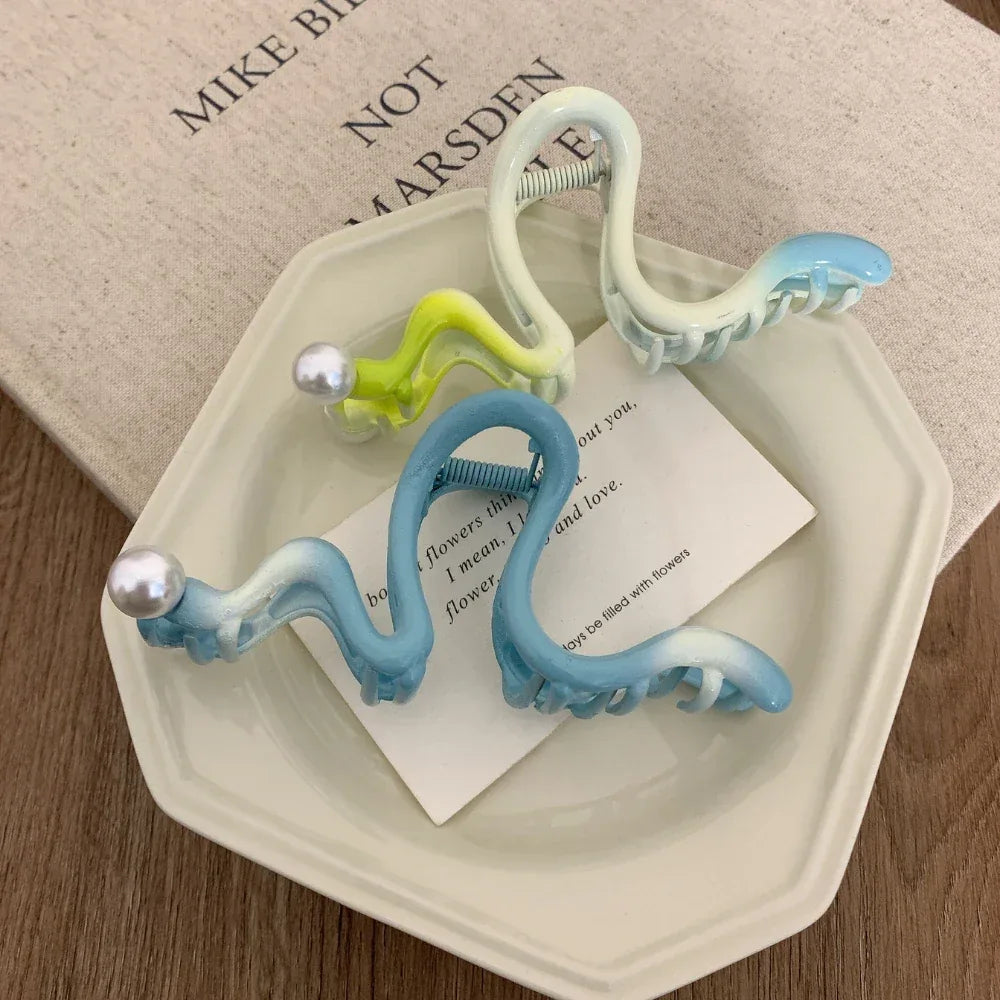 Close-up of light blue, green, and white S-shaped hair claw clips with pearls on a saucer