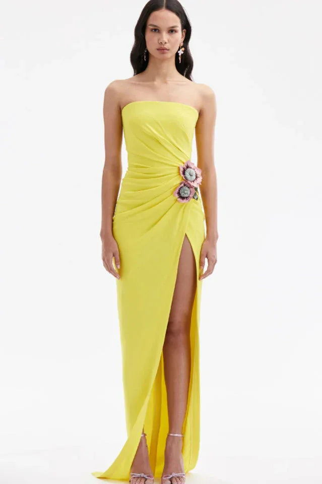 Full body shot of a woman in a yellow strapless gown with a high slit and floral embellishment against a white background.