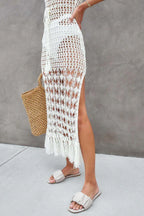 Close-up of the lower half of a woman's body wearing a white crochet knit maxi dress with a fringe hem and side slit, and sandals.